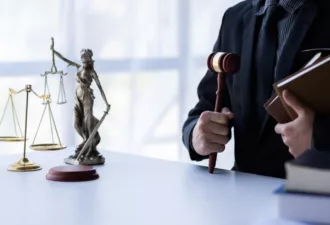 Un juge en costume noir tenant un marteau de justice et des livres de droit, avec une balance dorée et une statue de la justice sur un bureau blanc.